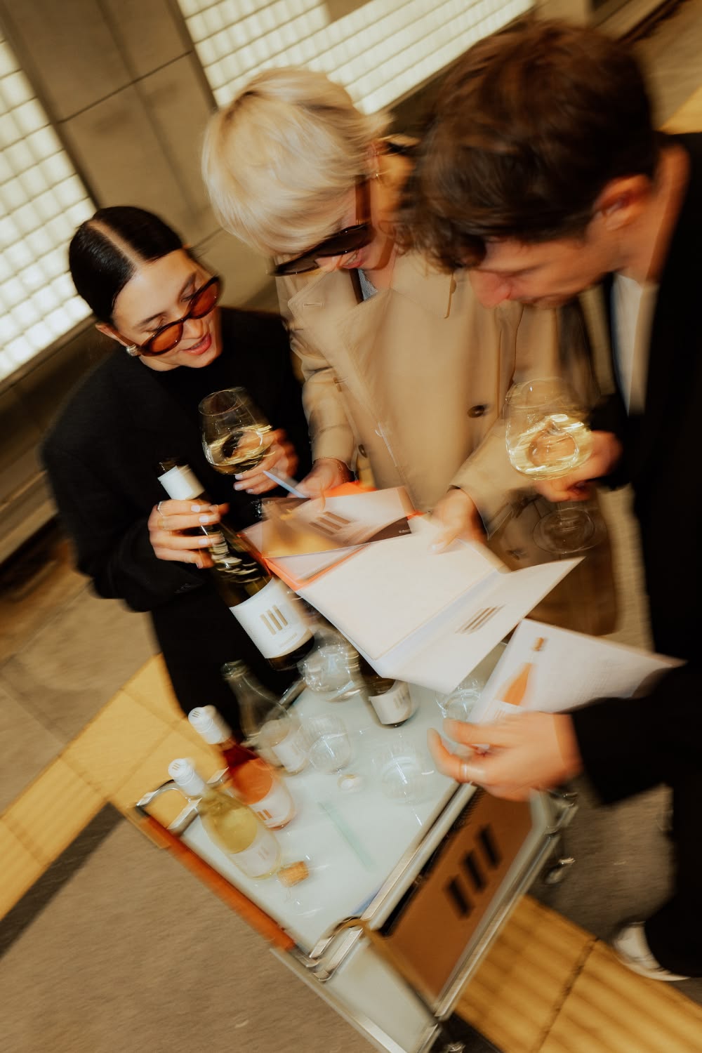Three colleagues with glasses of white wine reading tasting sheets over a cart of bottles